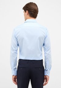 Camicia a maniche lunghe azzurra chiara con taglio dritto e polsini mancini, abbinata a pantaloni scuri con doppie tasche posteriori.
