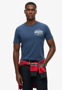 T-shirt bleu marine en coton, arborant un logo vintage en blanc. Stylisé avec un sweat à capuche rayé rouge et noir attaché autour de la taille.