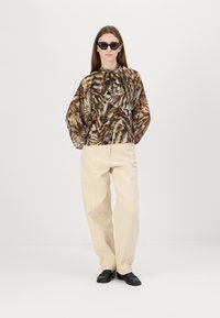 Brun og kremmønstret bluse med knytehals, kombinert med beige bukser og svarte loafers. Modellen har på seg svarte solbriller.