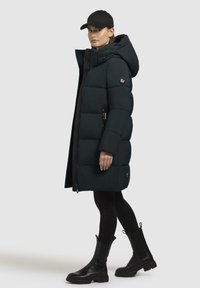 Geacă puffer neagră cu glugă, închidere cu fermoar și nasturi, design matlasat, buzunare laterale și patch circular cu logo. Purtați cu colanți negri și cizme.