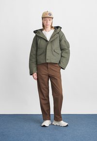 Olīvu zaļš kropts žakets ar lielām kabatām un aukliņu kapuci, uzvilkts virs baltas krekliņa un brūnām biksēm, apvienots ar baltām sporta kurpēm.