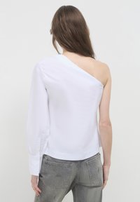 Eine Frau mit langen braunen Haaren trägt eine weiße asymmetrische Bluse mit einem Ärmel und ausgewaschene graue Jeans, während sie vor einem schlichten hellen Hintergrund steht.