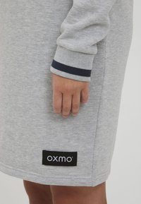 Szürke pulóverruha puha anyagból, lekerekített aljjal és bordás mandzsettával, amelyet egy sötétkék csík díszít. Tartalmaz egy fekete címkét az "OXMO" márkázással.