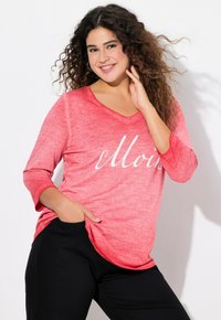 Maglia rossa melange a maniche lunghe con scollo a V e testo in bianco. Pantaloni neri con tasche completano l'outfit. Capelli ricci incorniciano il viso.