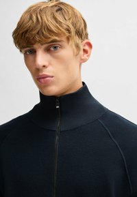 Maglione zip-up blu navy con un alto colletto a coste, morbido al tatto e dal design aderente con maniche raglan. La cerniera aggiunge un dettaglio funzionale.