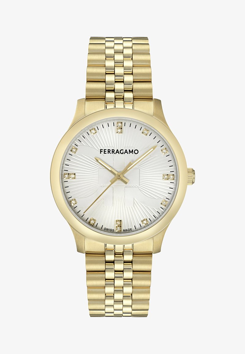 Arany színű óra ezüst, texturált számlappal, kristály órijelzőkkel és arany mutatókkal, arany láncos karkötőn. A logó hangsúlyosan megjelenik.