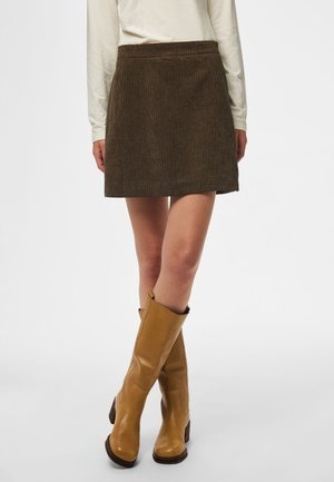 Femme portant une mini-jupe en velours côtelé marron, un haut beige à manches longues et des bottes en cuir marron clair montant jusqu'aux genoux, debout, jambes croisées.