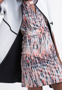 Robe ajustée colorée avec un motif ondulé en rose, bleu et noir, associée à un blazer cape blanc avec des bordures noires. Texture de tissu lisse.