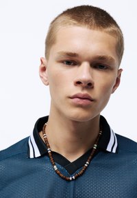 Manlig modell iförd en marinblå mesh-pikétröja med svarta krage och vita ränder. Han har ett halsband av träpärlor med färgglada inslag.