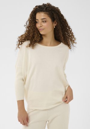 Donna con capelli ricci che indossa un top a maniche lunghe color crema e pantaloni abbinati, che guarda leggermente verso sinistra contro uno sfondo semplice.