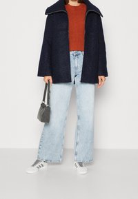 Laivansinisestä villasekoitekankaisesta takista, jossa on leveä kaulus, ruosteenvärisestä ribbikuvioidusta neulepaidasta, vaaleansinisistä leveälahkeisista farkuista ja harmaasta käsilaukusta. Valkoiset lenkkarit, joissa on harmaat yksityiskohdat.