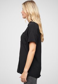 T-shirt nero a maniche corte con vestibilità rilassata, dotato di una tasca frontale e tessuto testurizzato. La vista laterale mostra un orlo posteriore leggermente più lungo.