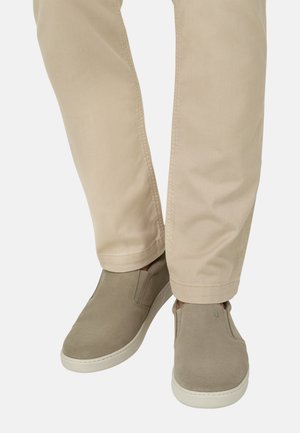 Voeten met beige slip-on suède schoenen met witte zolen en licht kaki broeken tegen een effen achtergrond.