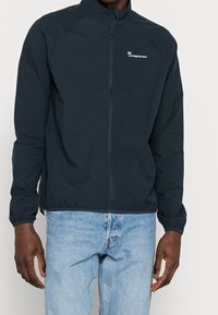 Námořnická větrovka s vysokým límcem, zipovým zapínáním a elastickými manžetami. Na levé hrudi se nachází bílé logo.