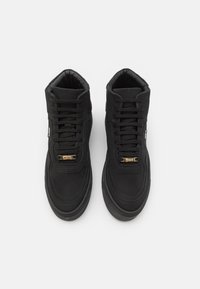 Para czarnych wysokich sneakersów z grubymi podeszwami, czarnymi sznurówkami oraz złotymi metkami z logo na białym tle.