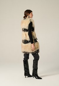 Gilet in pelliccia sintetica con un design a righe beige e nere, indossato sopra un top aderente nero e abbinato a stivali al ginocchio in vernice nera.
