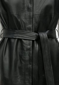 Fekete bőr ruhadarab, amely testhezálló kialakítású, gombos elülső résszel és kötött derékövvel rendelkezik. Simább textúrájú, látható varrási részletekkel.