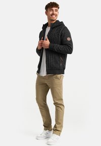 Czarna dziana hoodie na zamek z fakturowanym wzorem, z kapturem i stonowanymi akcentami. W połączeniu z beżowymi spodniami i białymi sneakersami.