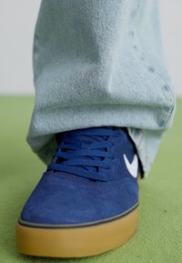 Baskets en suède bleu marine avec une semelle en caoutchouc beige, présentant des détails de logo blanc et des lacets plats, assorties à un pantalon en denim clair.
