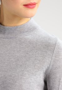 Ljust grått stickat tröja med hög ribbad krage och sömlösa axlar, som visar en mjuk, slät textur och minimala sömnadsdetaljer.