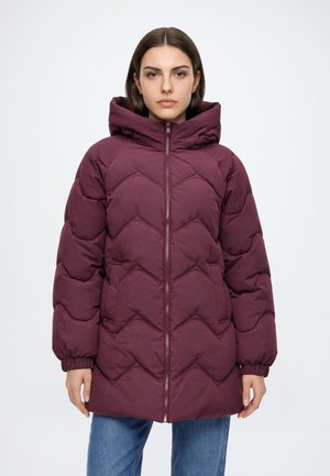 Mujer con una chaqueta de invierno larga acolchada de color granate con capucha, de pie con las manos relajadas a los lados contra un fondo claro y sencillo.