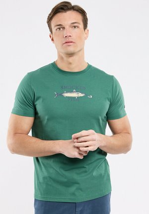 Homme à la peau claire et aux cheveux bruns portant un t-shirt vert avec un motif de poisson et le texte « SMOCKED FISH », les doigts entrelacés devant lui.