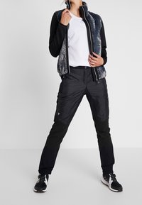 Chaleco negro con cremallera y forro de piel gris, combinado con pantalones cargo negros con paneles texturizados y bolsillos con cremallera, usado con zapatillas deportivas negras.