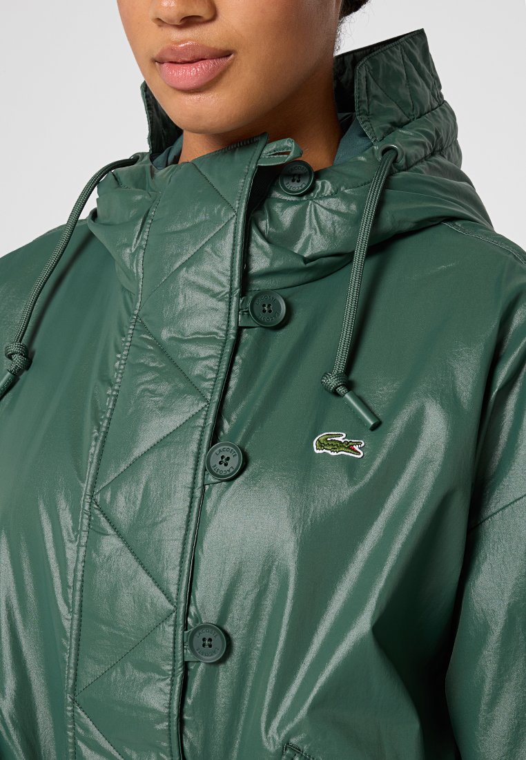 Chaqueta acolchada verde con un acabado brillante, que cuenta con capucha, cierres de botones y un pequeño logotipo bordado en el pecho.