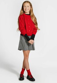 Czerwony, błyszczący kardigan z czarnymi mankietami, zestawiony z spódnicą w czarno-białą kratkę. Noszony z czerwonymi skarpetkami do kolan i czarnymi butami.