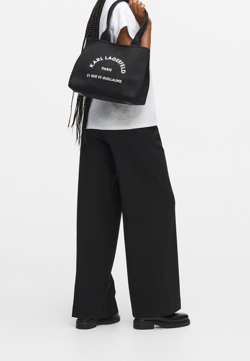 Personne portant un pantalon noir à jambes larges et une chemise blanche, tenant un sac cabas noir Karl Lagerfeld avec un texte blanc sur l'épaule.