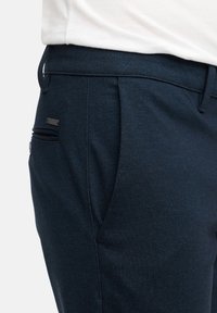 Marineblauwe herringbone broeken met een getailleerde pasvorm, voorzien van een retailtag en schuine zijzakken. Zachte textuur met gestructureerde ontwerpelementen.