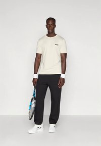 Mees, kellel on seljas valge BOSS t-särk, mustad püksid, valged tossud ja randmevalgustid, hoiab käes sinist ja musta tennisereketit, seistes puhta tausta ees.