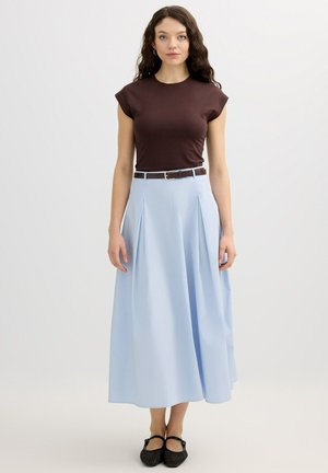 Joven con cabello largo, oscuro y rizado, vestida con un top ajustado marrón oscuro, falda midi plisada azul claro, zapatillas negras y cinturón marrón, de pie.