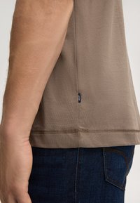 Korte mouwen, taupe t-shirt van zachte katoen. Heeft een rechte zoom met contrasterende stiksels en een klein zwart merkje aan de zijkant.