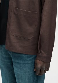 Zbliżenie osoby w brązowej kurtce z przednią kieszenią i niebieskich dżinsach, pokazującej prawą rękę spoczywającą przy boku.