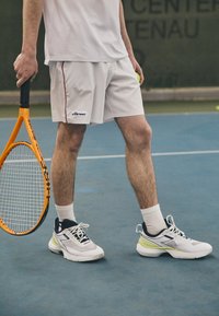 Weiße Sportshorts mit roten Akzenten, ein weißes Mesh-Oberteil und schwarz-weiße Tennisschuhe mit limettengrünen Details. Hält einen orangefarbenen Tennisschläger.