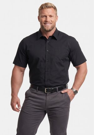Smilende mand med kort blondt hår iført en sort kortærmet skjorte med knapper, grå bukser, sort bælte og sølvfarvet armbåndsur.