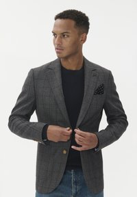 Moški zapenja gumb na sivo karirastem blazerju čez črn pulover, v modrih kavbojkah, z črno pikasto maramico v žepu, gleda levo.