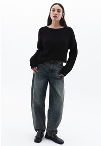 Maglione nero a coste, jeans in denim blu sbiadito a taglio comodo e mocassini neri. Appare informale e rilassato.
