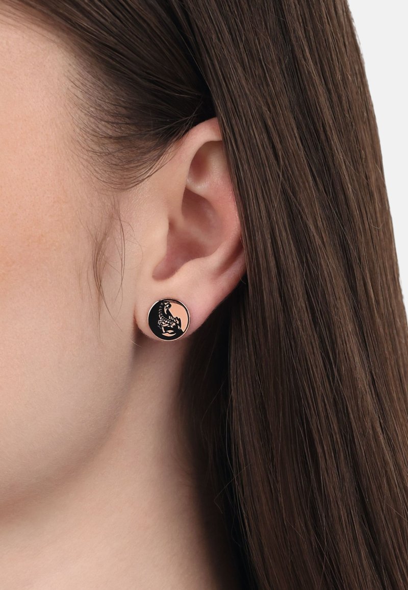 Zwarte stud oorbellen met een gedetailleerd ontwerp van een zittende figuur, gezet tegen een ronde, blush achtergrond. Gladde, metallic textuur.