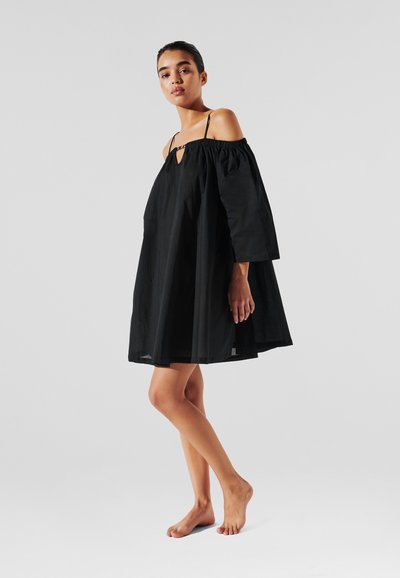 Robe noire à épaule dénudée avec des manches trois-quarts, en tissu léger, un détail en forme de trou de serrure au niveau du col, et une silhouette ample et fluide.