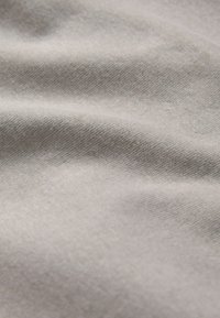 Nahaufnahme eines weichen, hellgrauen Stoffes mit feiner, diagonaler Stricktextur und sanften Falten, die subtile Schatten erzeugen.