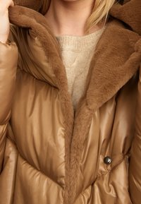 Bruine puffer jas met een zachte, nepbont voering. Het ontwerp heeft een brede kraag en knopenaccenten, waarmee de textuur van de jas wordt benadrukt.