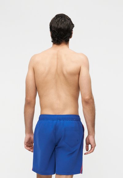 Voksen mann sett bakfra, uten skjorte, iført blå badetruser med baklomme mot en enkel lys bakgrunn.