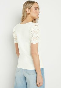 T-shirt blanc léger avec des manches bouffantes en crochet, design ajusté et texture lisse. La vue de dos montre une couture subtile et un col rond.