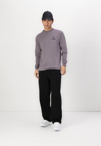 Grå langermet skjorte med en liten svart trekantlogo. Modellen har på seg svarte vide bukser og hvite joggesko, toppet med en svart caps.