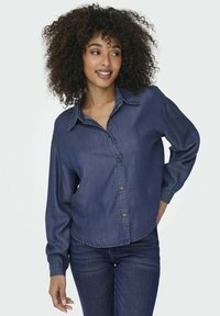 Camicia in denim blu scuro con bottoni, dotata di colletto aperto, maniche lunghe e orlo curvo, con una texture morbida e una vestibilità standard.