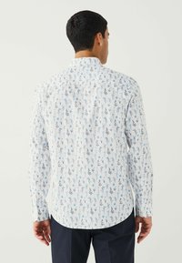 Bărbat îmbrăcat cu o cămașă albă cu mâneci lungi, având un mic model floral albastru, întors cu spatele, asociată cu pantaloni închiși la culoare, pe un fundal simplu de culoare deschisă.