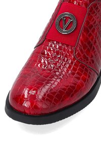 Czerwone lakierowane buty z fakturowanym wzorem krokodyla, zaokrąglony czubek, czarna podeszwa i wyraźne okrągłe srebrne logo na czerwonej wstążce.