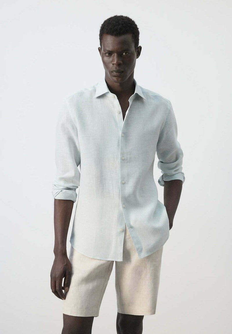 Homme portant une chemise légère à manches longues avec les poignets retroussés et un short beige sur un fond blanc uni.